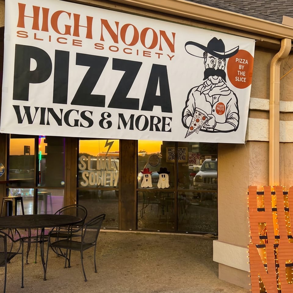High Noon Slice Society storefront with pizza banner, outdoor seating, illuminated OPEN sign, and sunset reflections in the windows.