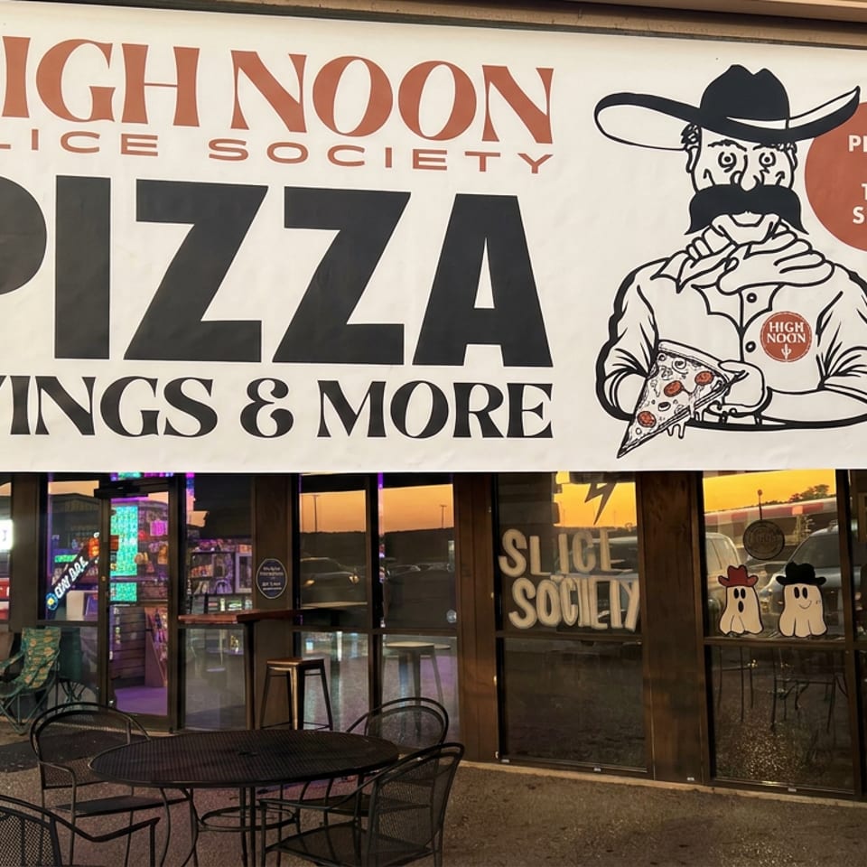 High Noon Slice Society storefront with a large pizza banner, cowboy mascot, outdoor tables, and glass entrance.