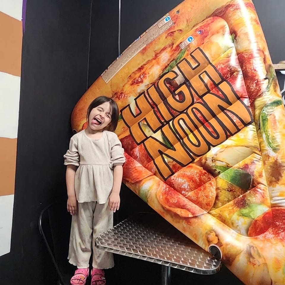 Child stands beside a giant colorful pizza slice prop reading “High Noon,” next to a “First 80” photo display.